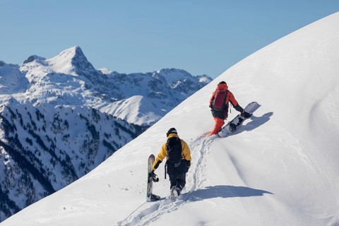 Two snowboarder friends trek a snowy mountain slope under clear blue skies, with majestic peaks in