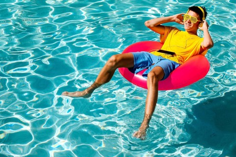 A young teenage boy in a yellow t-shirt and blue shorts relaxes on a red pool float while listening