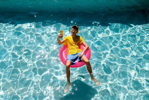 A teenage boy in a yellow shirt and shorts lounges on a pink float in a sparkling blue pool. They