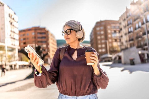 Woman with grey hair wearing headphones and stylish glasses, holding a coffee cup and smartphone