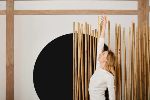 A woman stands in a room reaching her arm upward. She is positioned near bamboo sticks that are