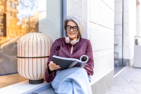 Mature woman with gray hair and glasses smiling gently while sitting on a city street sill,