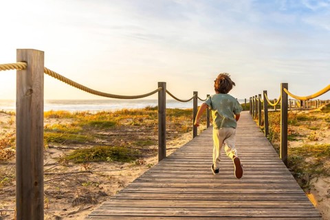 Little boy joyfully running along a wooden boardwalk towards the ocean as the sun sets, symbolizing