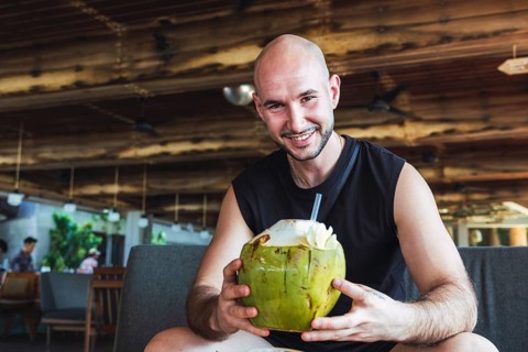 A cheerful bald man enjoys drinking a fresh coconut while relaxing in a rustic tropical cafe