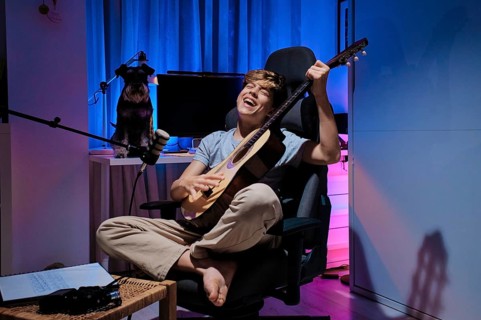 A joyful musician plays an acoustic guitar in a cozy, colorful home studio. A dog watches nearby,