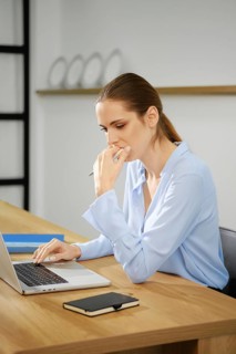 A young woman dressed in a light blue shirt is focused on her laptop while sitting at a wooden desk