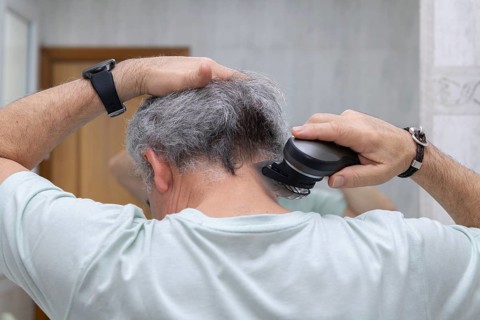 A man uses a hair clipper to trim the hair on the back of his head. He stands in front of a