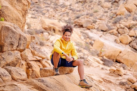 A young hiker in a yellow jacket sits on rugged rock formations of the Canary Islands, smiling