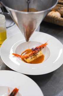 A chef precisely pours seafood sauce over a gourmet shrimp and terrine dish using a funnel,
