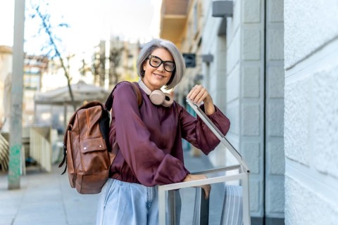 Woman with gray hair, glasses, and headphones wearing a backpack, smiling while pulling a piece of
