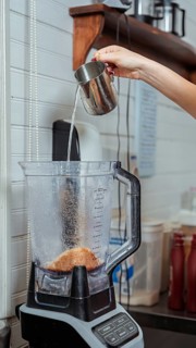 Hand pouring water from a metal pitcher into a blender filled with coffee grounds in a cozy cafe.