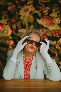 Chic grandma poses playfully in a studio setting, wearing sunglasses and gloves against a floral