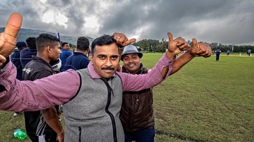 Two men give thumbs up on a sports field with cloudy skies, people and fans playing sports in