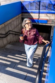 Woman with gray hair and glasses ascending subway stairs, holding a disposable coffee cup and