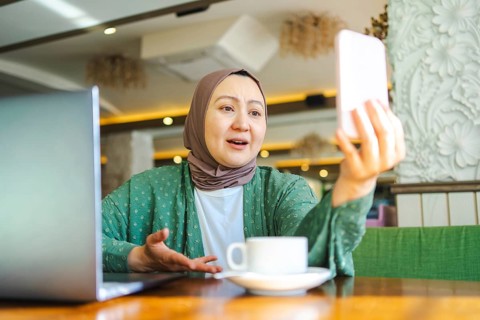 A Muslim woman in a hijab sits at a cafe table, engaging in a video call on her smartphone. A