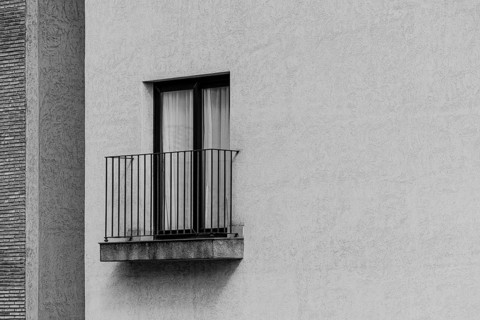 Black and white image of a minimalist exterior wall with a small balcony and a window. The textured