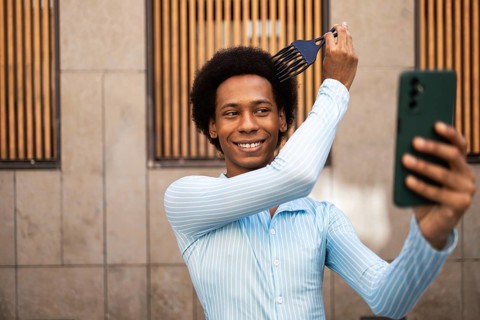 Smiling young non-binary adult styling hair with a pick comb while capturing a selfie. Wearing a