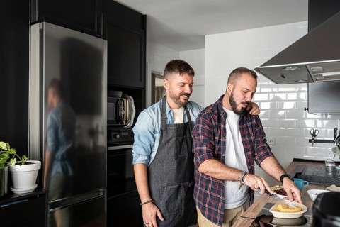 A loving gay couple prepares a meal together in their cozy, modern kitchen. One man embraces the