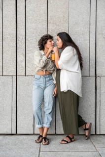 A lesbian couple shares a drink while leaning against a stylish concrete wall. They are enjoying a