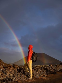 A person in a vivid red raincoat stands on volcanic terrain, gazing at a vibrant rainbow in