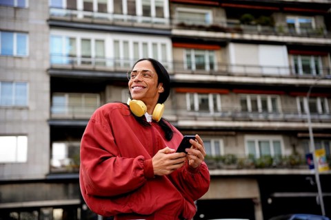 A man stands outdoors in a city environment, smiling while holding a smartphone. He wears glasses,