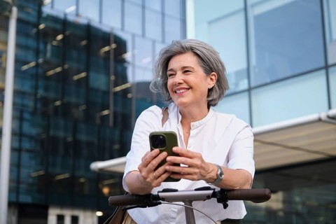 Mature woman smiling while holding a smartphone outdoors, dressed in a casual white shirt, enjoying