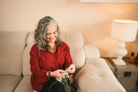 A woman with curly gray hair, wearing a red blouse, is knitting on a comfortable beige couch. A