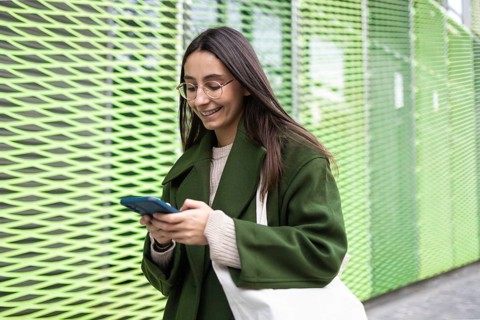 A young woman dressed in a stylish green coat is smiling while using her smartphone She stands