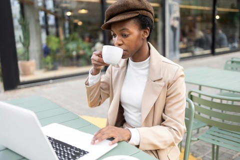 A black woman enjoys a cup of coffee while browsing on a laptop at an outdoor urban cafe She wears