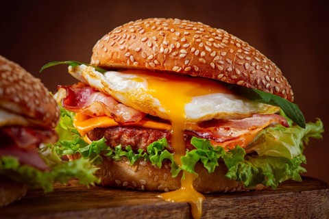 Two juicy burgers sit on a wooden board, each filled with lettuce, bacon, cheese, and fried eggs