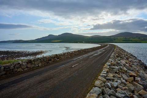 A rocky road stretches through a seascape with twilight sky and mountains in the background, Tongue