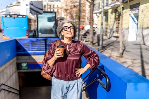 Senior woman with gray hair and glasses stands at a subway entrance holding coffee, looking up at