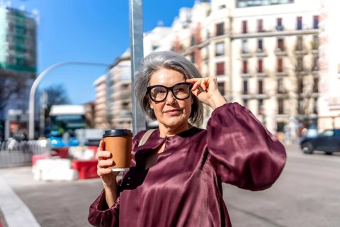 Mature woman with gray hair wearing glasses and a purple blouse, holding a disposable coffee cup