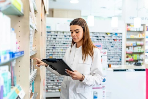 Female pharmacist wearing a white coat, holding a clipboard and pen, checking medication inventory
