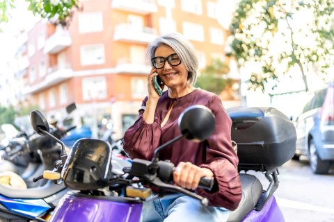 Mature woman on a scooter in a bustling city street, smiling and talking on her smartphone while