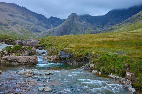 River flows through green valley plain in a mountainous landscape under cloudy sky, Fairy Pools,
