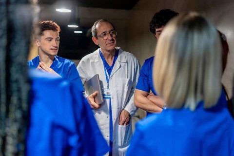 Experienced male doctor wearing a lab coat and holding a laptop, engaging with a group of young