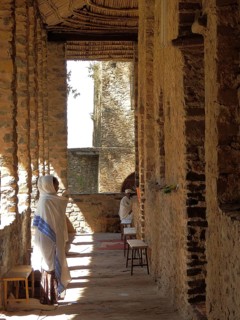 People wearing traditional clothing stand in a shady stone corridor, Debre Birhan Selassie Church,