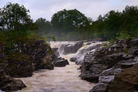A thundering river cuts through rocky formations surrounded by green vegetation and overcast sky,