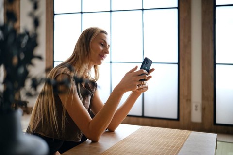 A woman sits at a wooden table in a room with large windows. She is focused on her phone, smiling