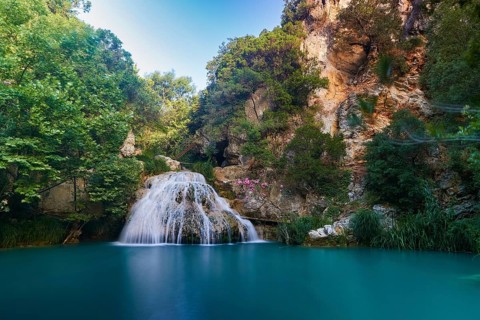 A waterfall falls into a turquoise-blue basin surrounded by green trees and rocky cliffs,