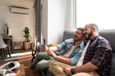 A joyful gay couple takes a selfie on a cozy couch in their modern living room, which features