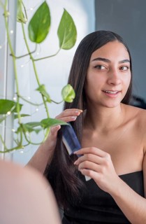 A woman with long, straight hair stands near a window, using a blue comb. She is smiling naturally,