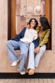A joyful lesbian couple in casual clothes sits by a shop window, sharing a light-hearted moment.
