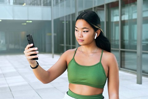 A young Chinese teenager holds a smartphone, wearing wireless earbuds. She is outside an urban