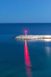 A peaceful scene of the Mediterranean Sea at dusk featuring a solitary lighthouse on a rocky pier.