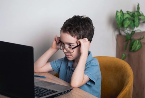 A young child in a blue shirt focuses intently on a laptop screen while adjusting glasses, seated