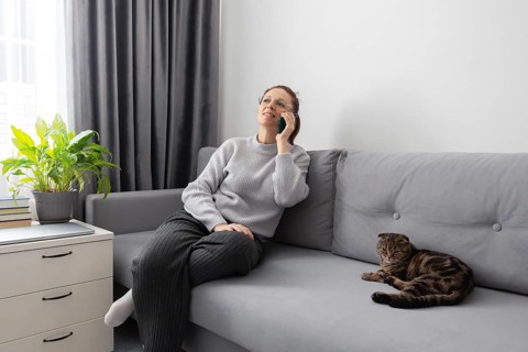 A woman sits on a gray sofa, talking on the phone, with a relaxed cat beside her. The room features
