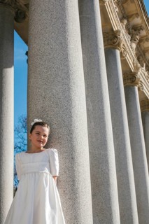 A young girl wearing a First Communion dress stands elegantly beside large stone columns, basking