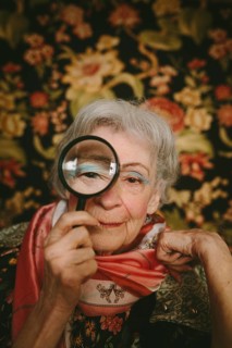 An elderly woman holds a magnifying glass to her eye, set against a vibrant floral backdrop. Her